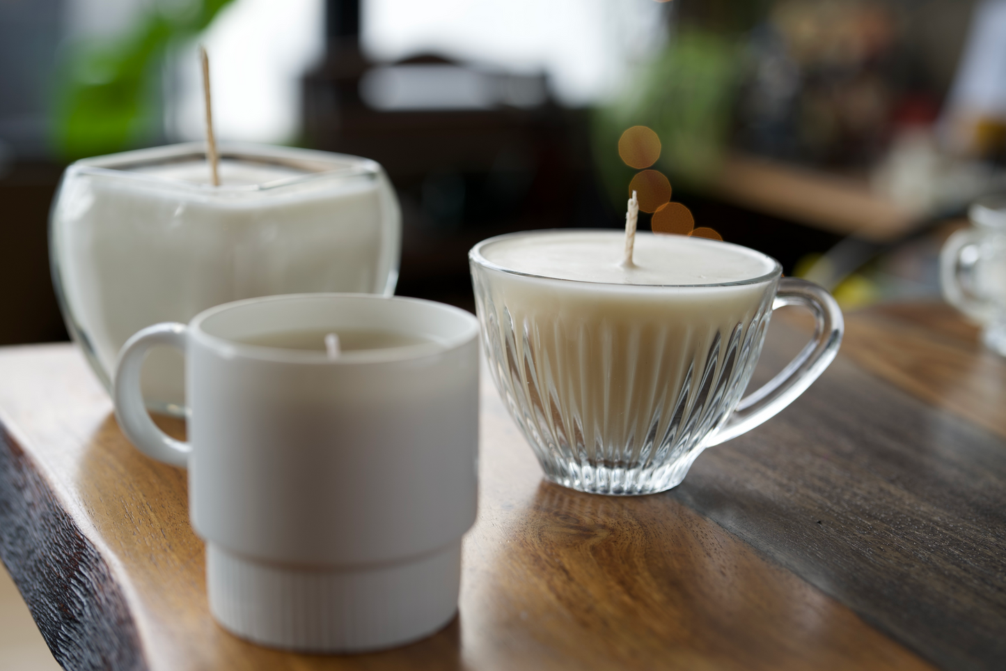 Three pure white, unlit soy wax candles. They are resting on a dark wood table, with bokeh lights in the background. One is white and two are clear glass. All three candles are modern, classy, and elegant.